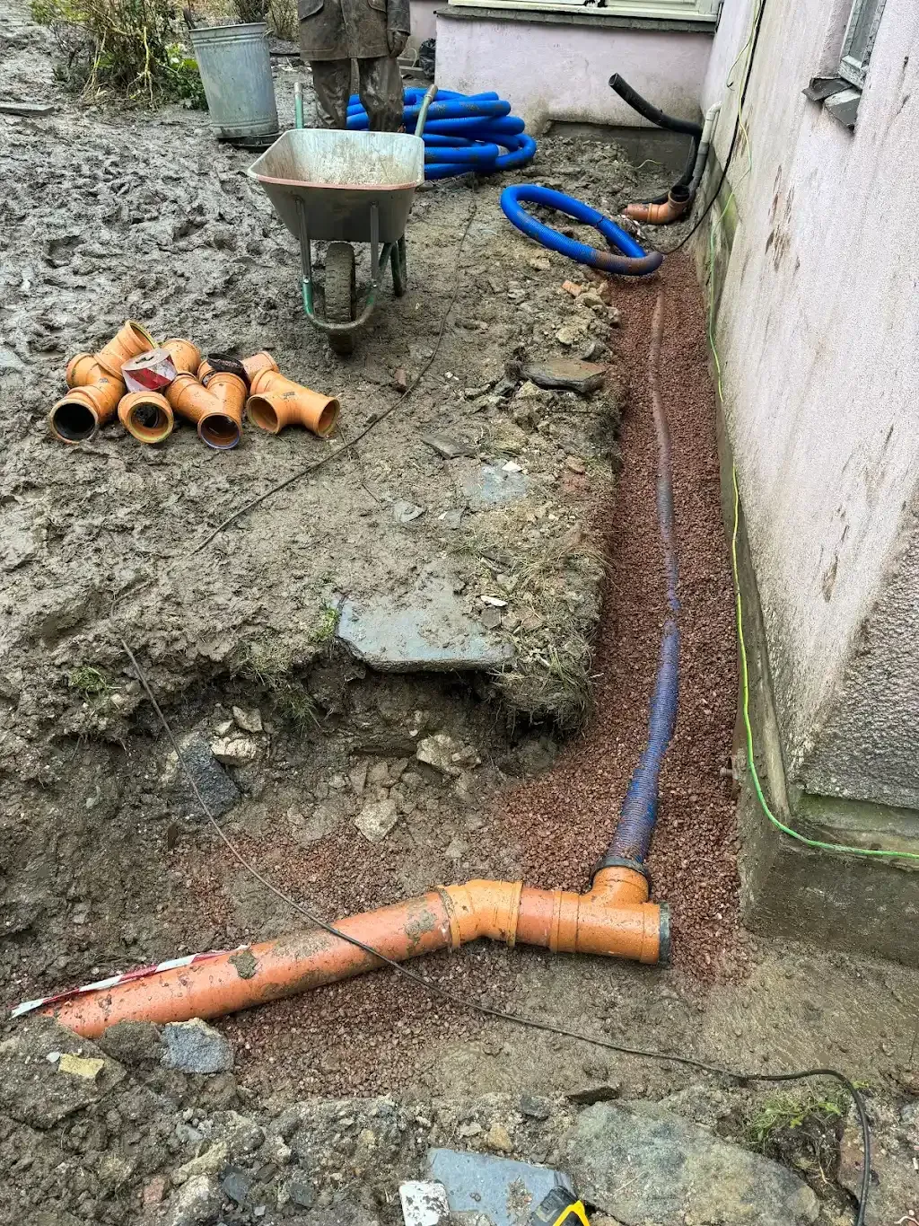 A trench with orange drainage pipes being installed next to a building foundation in Devon.